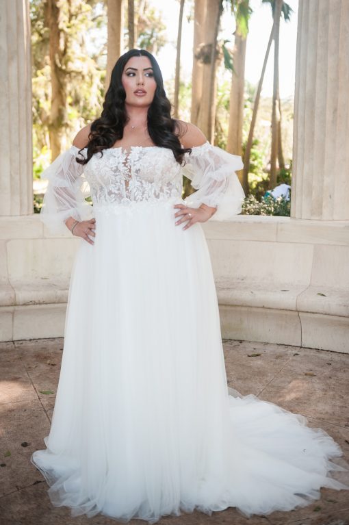 Plus‑size bride modeling Stella York 7573+; boho A‑line wedding gown with floral lace bodice, sheer detachable off‑shoulder tulle sleeves, plunging neckline, and soft flowing skirt.