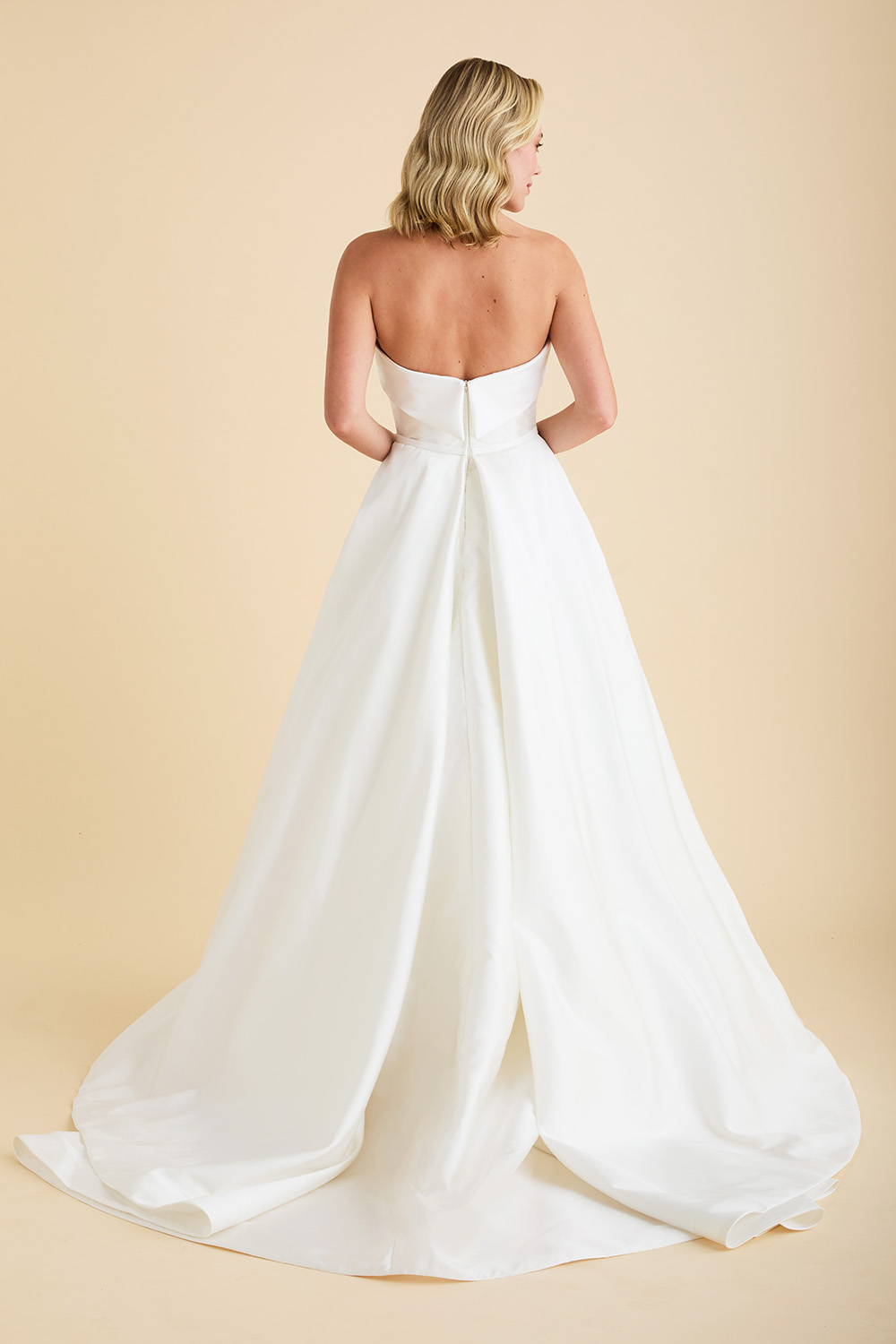 Off‑white Astrid & Mercedes Luster wedding dress in eco‑mikado, featuring a strapless straight‑across neckline with duchess satin band, tailored bodice, full skirt with pockets, chapel train, and inverted pleat