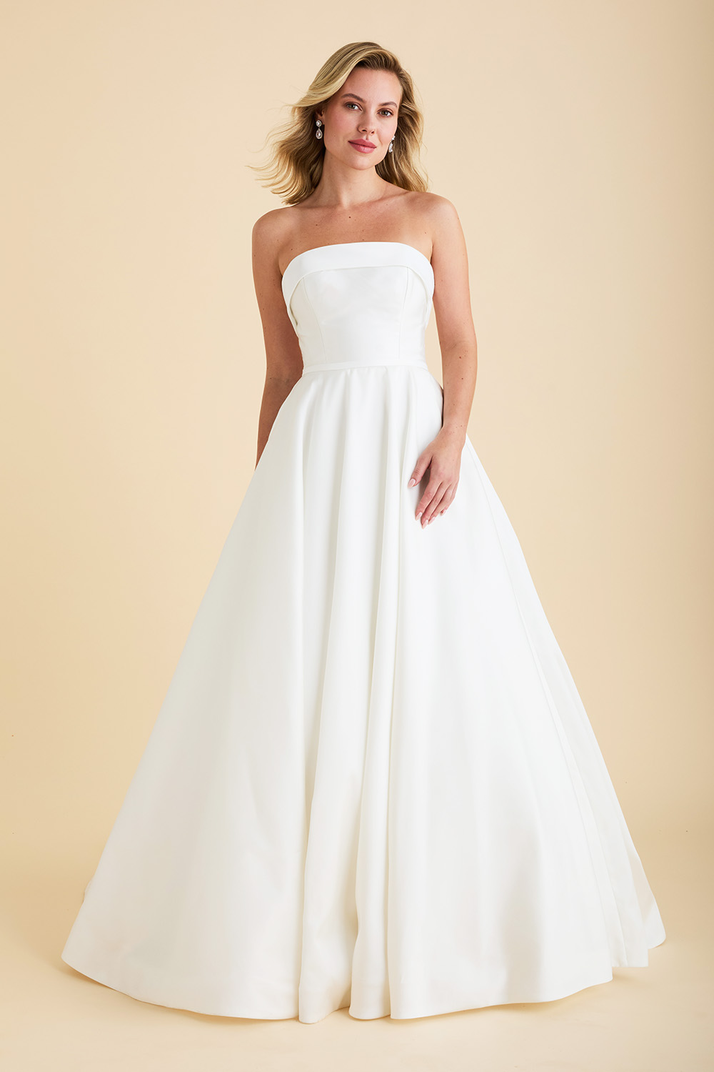 Off‑white Astrid & Mercedes Luster wedding dress in eco‑mikado, featuring a strapless straight‑across neckline with duchess satin band, tailored bodice, full skirt with pockets, chapel train, and inverted pleat